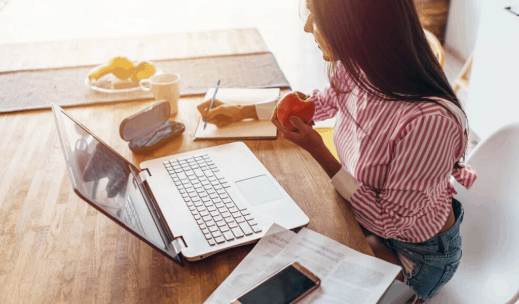 woman at desk looking at computer taking notes eating an apple