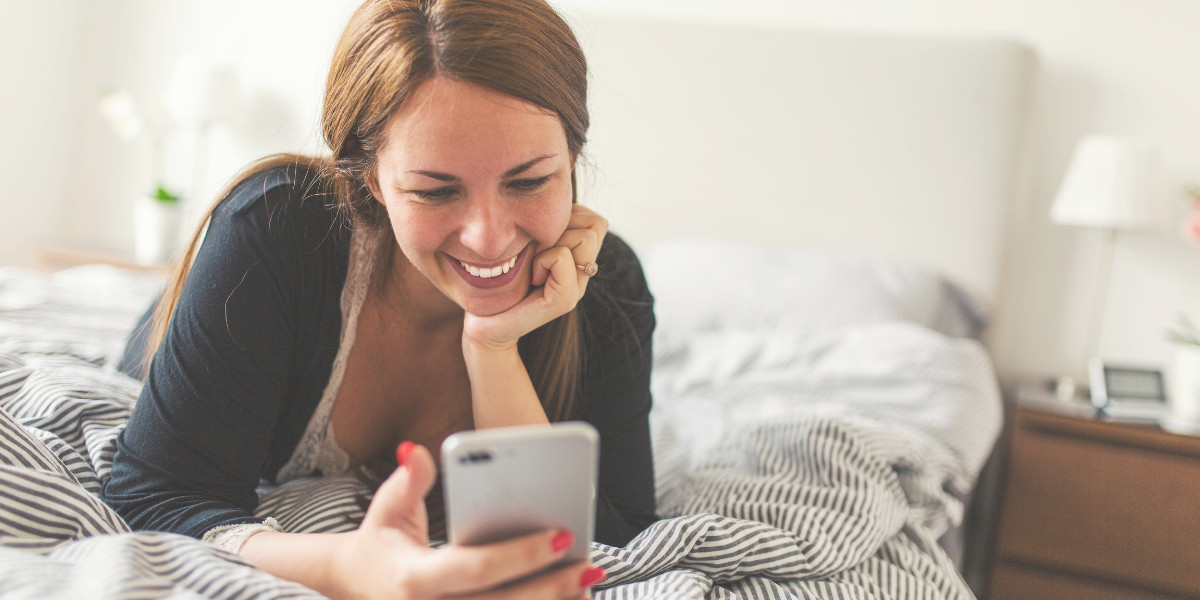 White woman laying on a bed searching on her hone for the best money making apps for 2020.