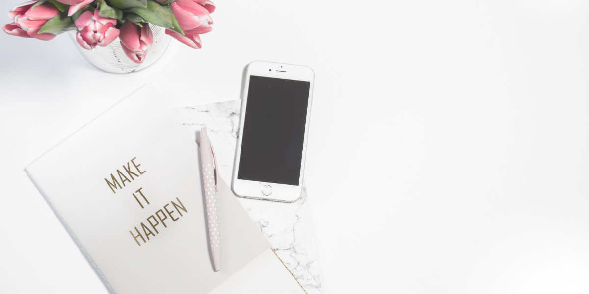 "Make it Happen" Notebook on a desk with a cellphone and flowers next to it