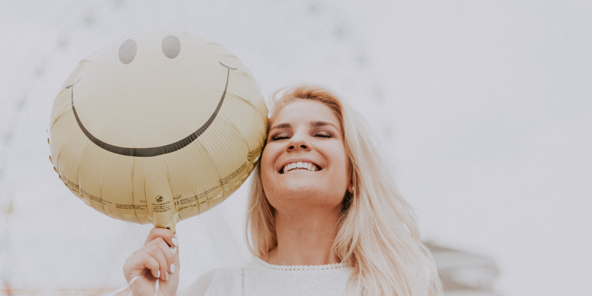 White female smiling while holding a yellow balloon with a smiley face 