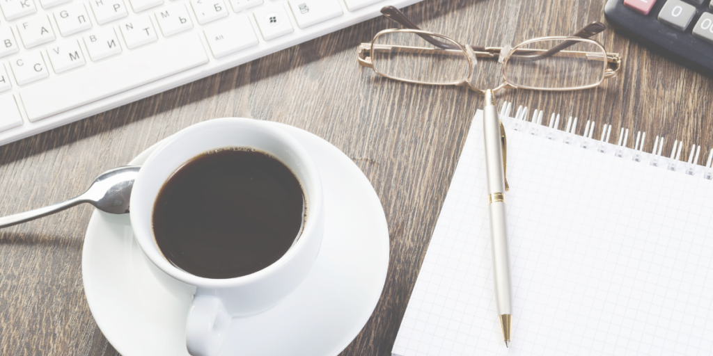 Cup of coffee sitting on a desk with a pair of glasses and pen and notebook sitting next to it