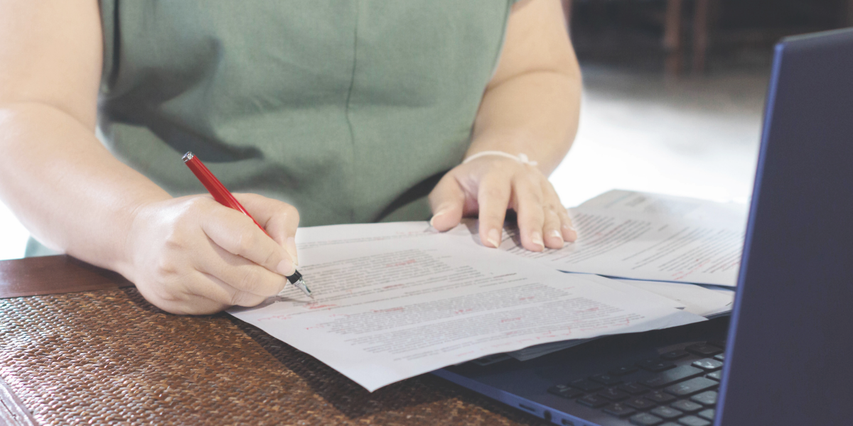 White female proofreading a document. Laptop sits next to her on the desk.