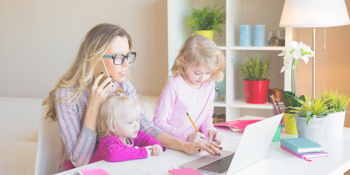 Mother working on her laptop with one small daughter in her lap and another daughter standing next to her drawing on a sheet of paper