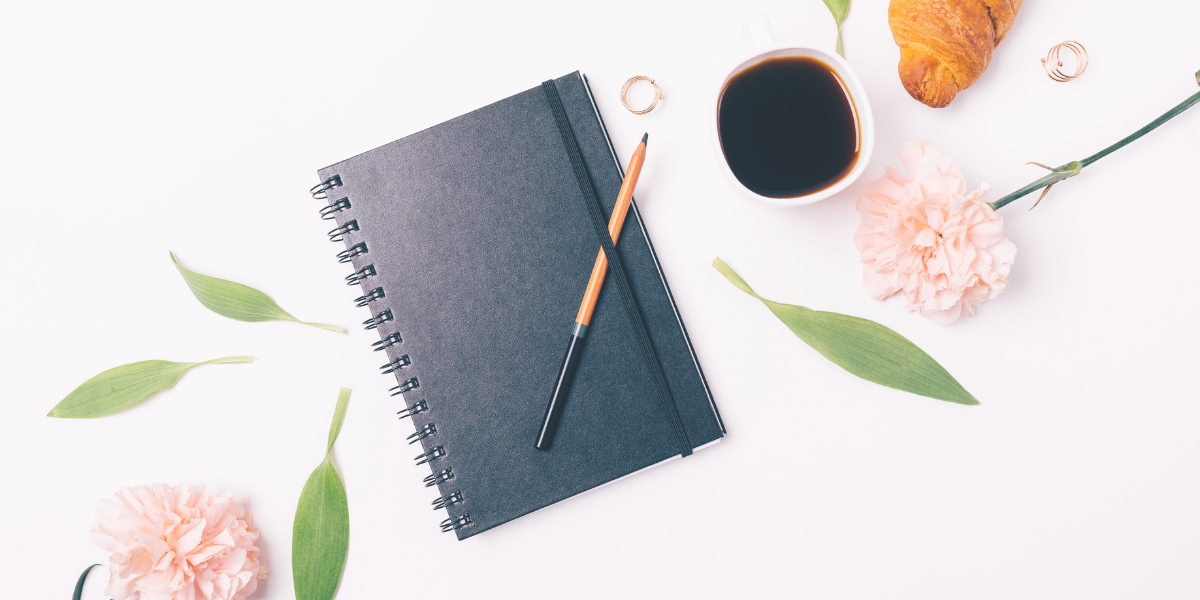 Black notebook lying on a desk with a cup of coffee sitting beside it as well as some pink flowers.