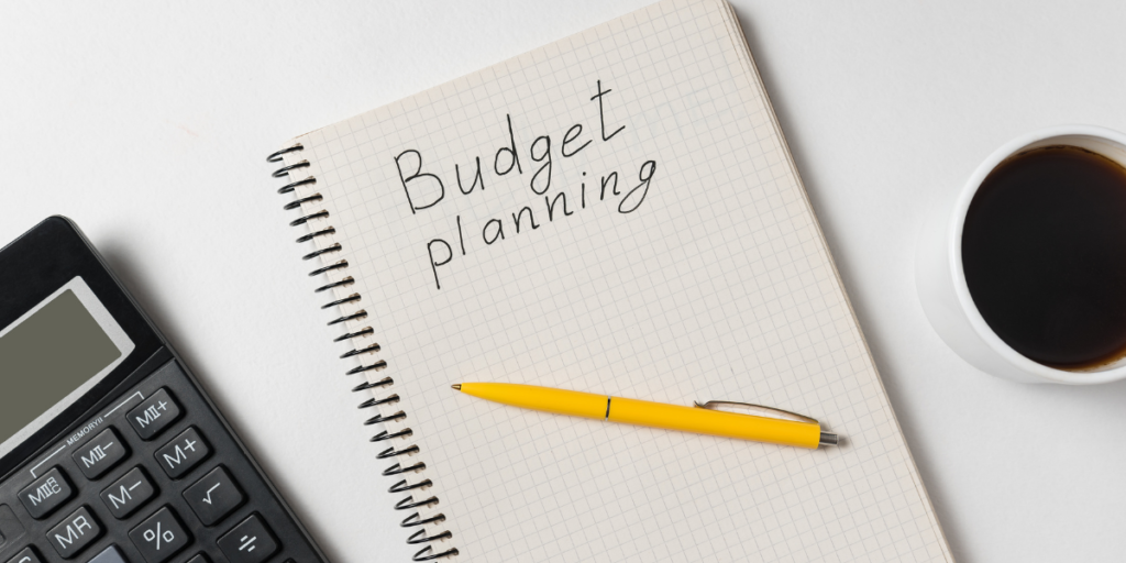 A white notebook with a yellow pen, calculator and coffee on a white desk  with budget planning written on the page with plans to change money mindset and secure a financial future. 