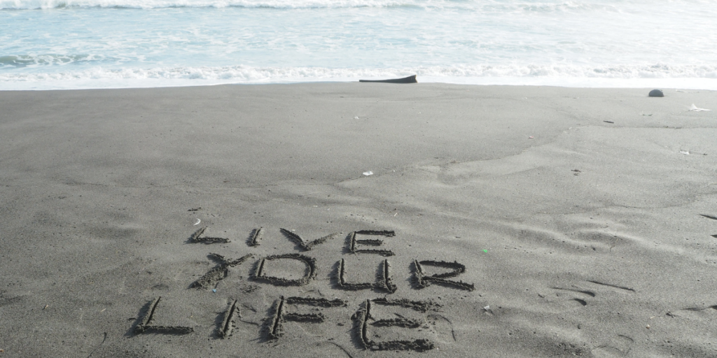 Live Your Life written in the sand on the beach reminding us to not worry about keeping up with the Jones', but to worry about our own financial future and changing our money mindset to reach our own goals. 