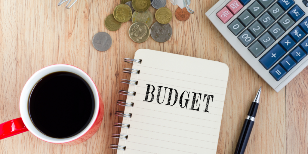 A notebook laying on a table beside a cup of coffee, a pen, a calculator and some change that is being used to stick with a budget. 