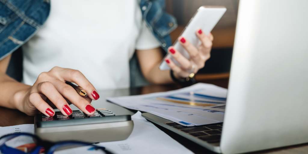 A woman sitting in front a laptop covered in paperwork, using a calculator and looking at her cellphone while working on reviewing and sticking to her budget. 