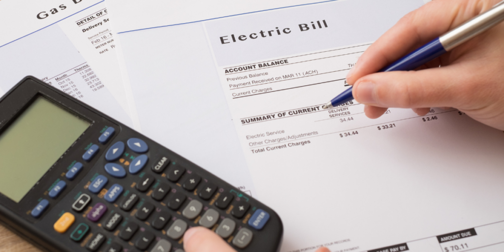 A calculator laying on top of the electric and gas bills with a person holding a pen reviewing the bills in an effort to stick to their budget. 