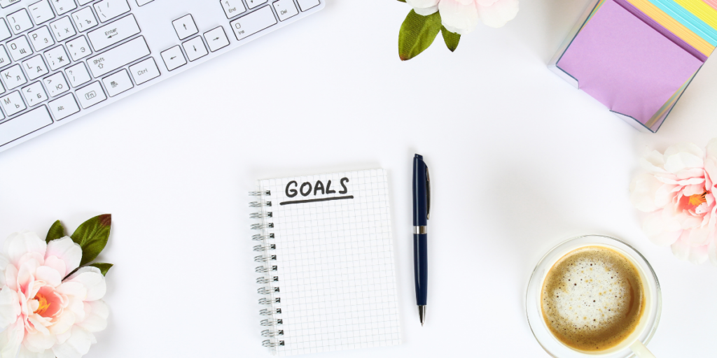 A desk with a keyboard, post it notes with a cup of coffee next to a notebook with goals written across the top of the paper and a pen next to it used to write down ones financial goals. 