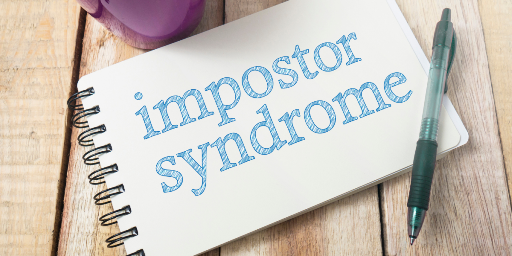 A notebook with impostor syndrome printed on the page with a coffee cup and pen nearby to be used to stick to a budget. 