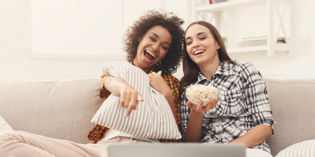 Two friends sitting on a couch watching a free movie online using a laptop and eating popcorn. 