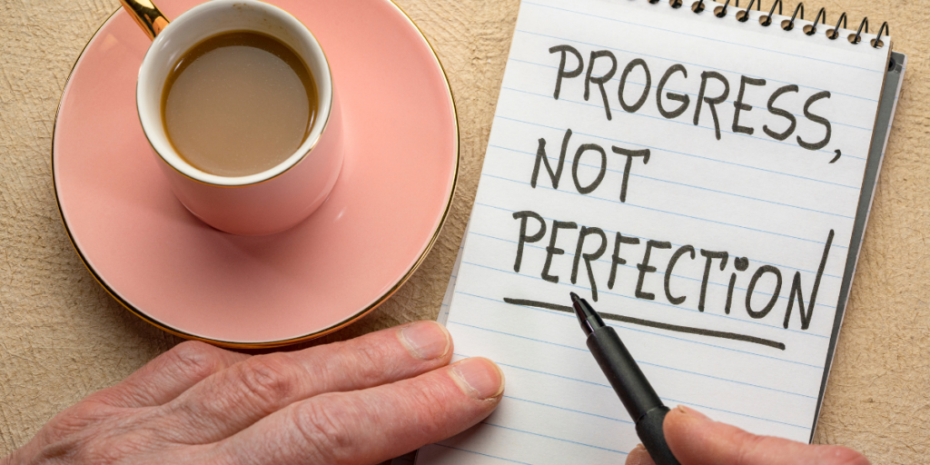 A person enjoying a cup of coffee with a pen and notepad that says "Progress, Not Perfection" reminding us progress is needed in order to reach financial goals. 