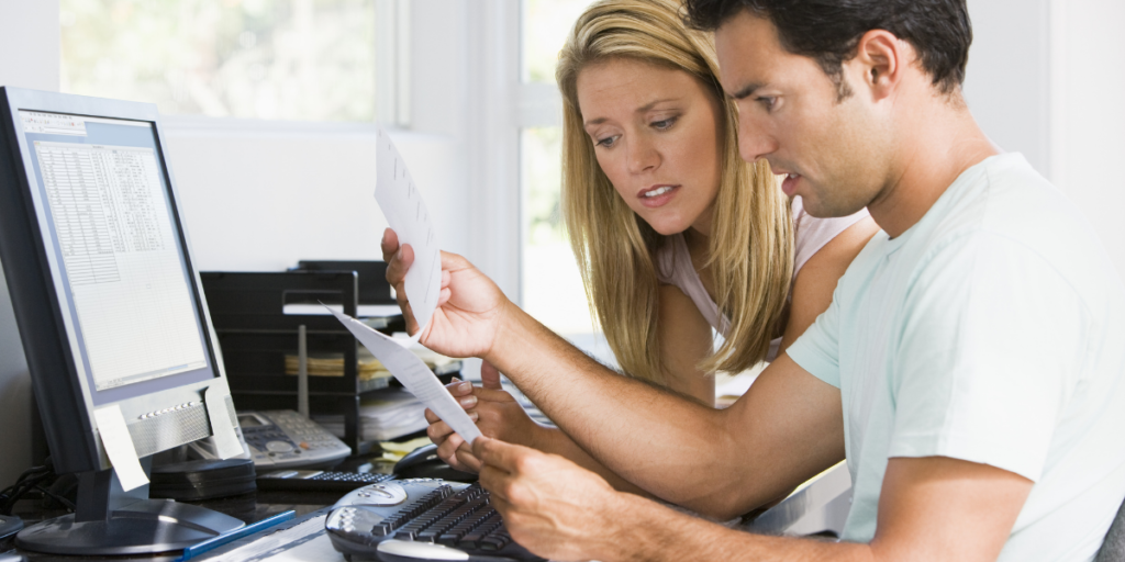 A man and a woman reviewing paperwork in front of a computer sitting at a desk discussing sticking to their budget. 