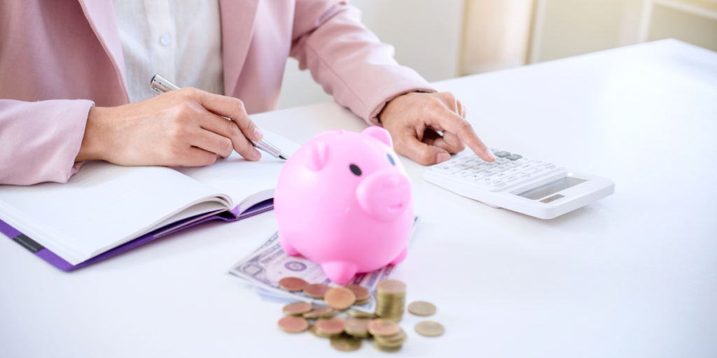 A woman sitting at a desk with her notebook, pen, calculator and piggy bank with money laying out while she's working on sticking to her budget. 