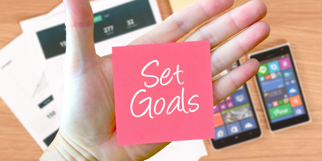 Cellphones and paperwork laying on a desk with a hand hovering above holding a dark pink post-it note with the words "set goals" in white reminding one why they should stick to a budget. 