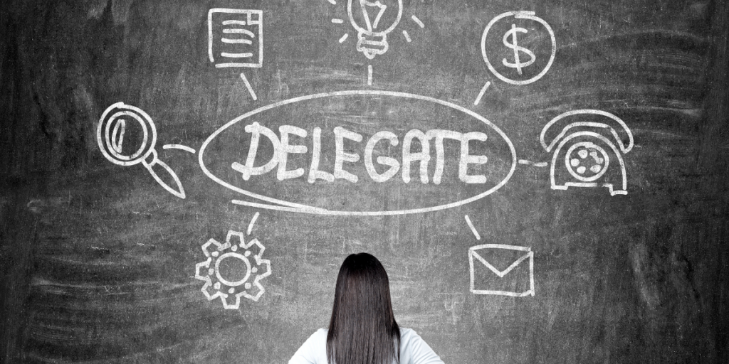 A chalkboard with the word delegate written on it as a female looks at items drawn around the work that she can think about delegating to other to help her stay motivated. 