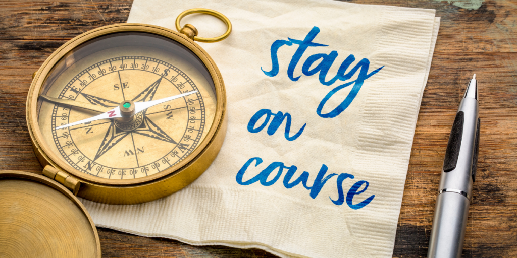 A compass laying on a napkin with the words stay the course on it with a pen nearby and remember to stay motivated. 