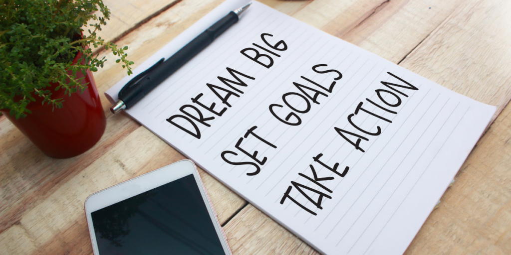 Staying motivated means reminding yourself to Dream Big Set Goals Take Action written on a piece of paper with a pen sitting at the top of the paper and a cellphone near by.