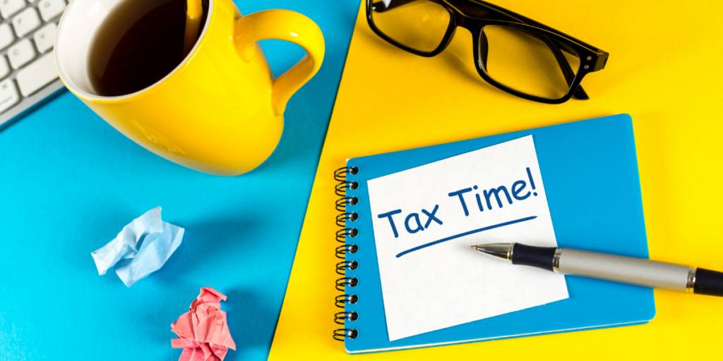 A tax time post it note laying under a pen on a table by a laptop, eye glasses, crumpled paper and a large cup of coffee. 