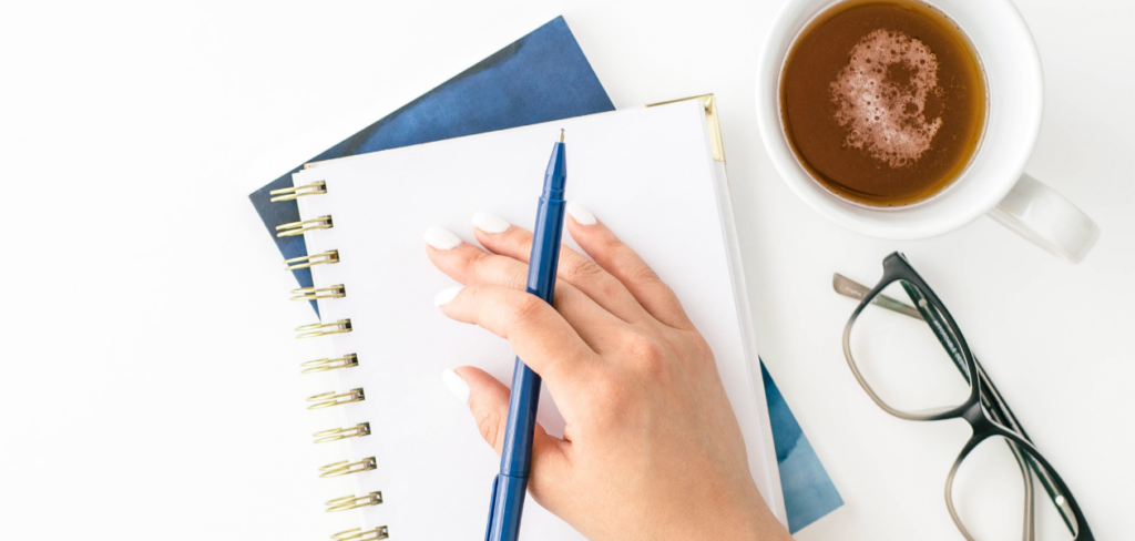 a hand holding a blue pen with a notebook underneath and a cup of coffee beside a reading glasses