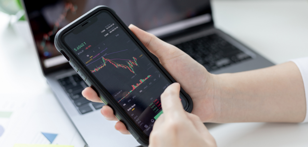 a hand holding a phone showing the stock market with a laptop at the back