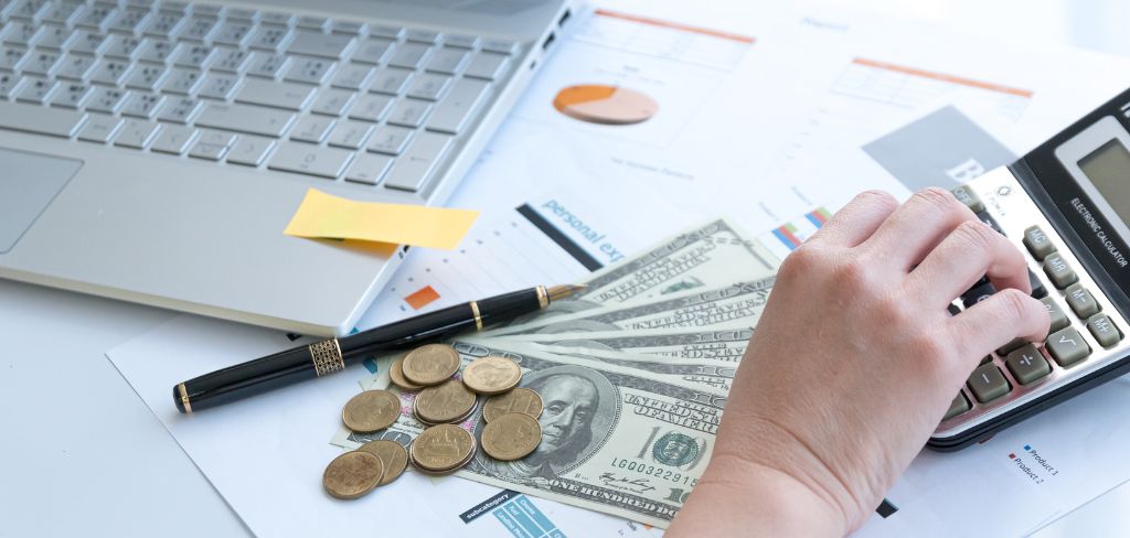 Hand calculating on calculator, money and coins, laptop and pin on desktop.