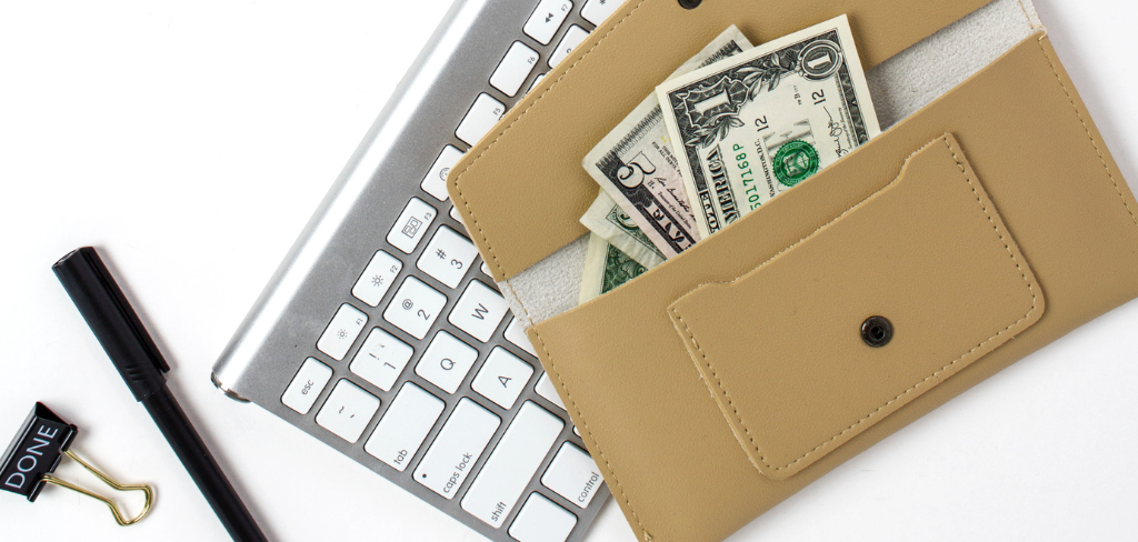 Keyboard with black ink pen and black clip and a beige wallet with five and one dollar bills.