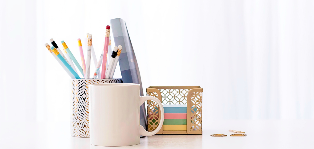 Empty white coffee cup, coffee cup with pencils and pens, planner and gold post it note holder with post it notes.