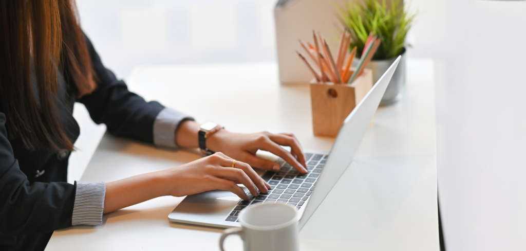 Image of a woman hands typing on a computer laptop on a white working desk surrounded by a coffee mug.