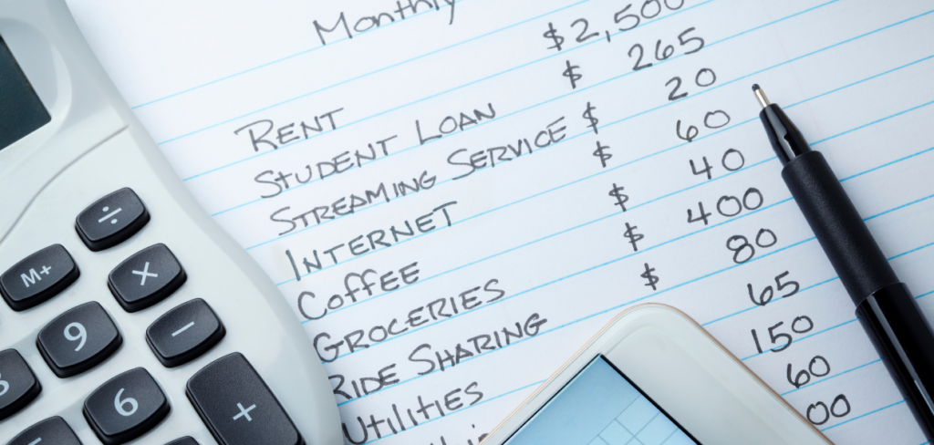 A calculator and a mobile device rest on top of a list of monthly expenses written on a sheet of paper.