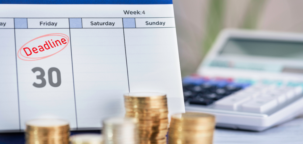 A desk calendar with a deadline, stacks of coins, and calculator on a desk.