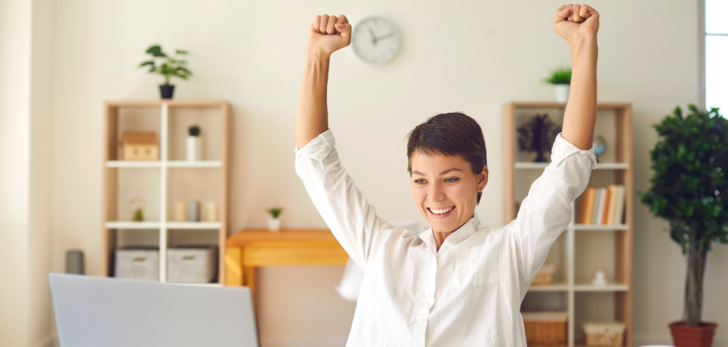 A woman is sitting at the table in front of a laptop, looking joyful as she raises her hands in celebration.