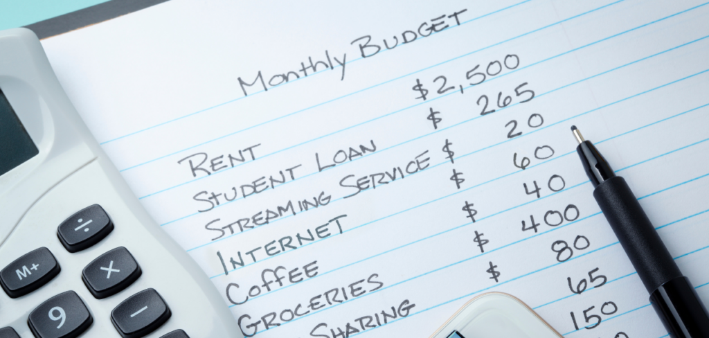 A calculator and a mobile device rest on top of a list of monthly expenses written on a sheet of paper.