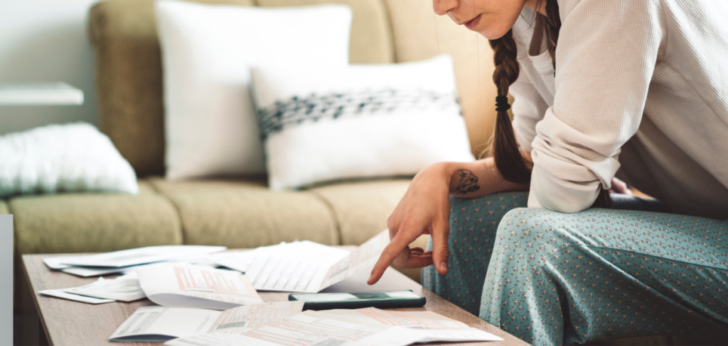 A woman is sitting on the sofa with bills on the table, checking her finances on her smartphone.
