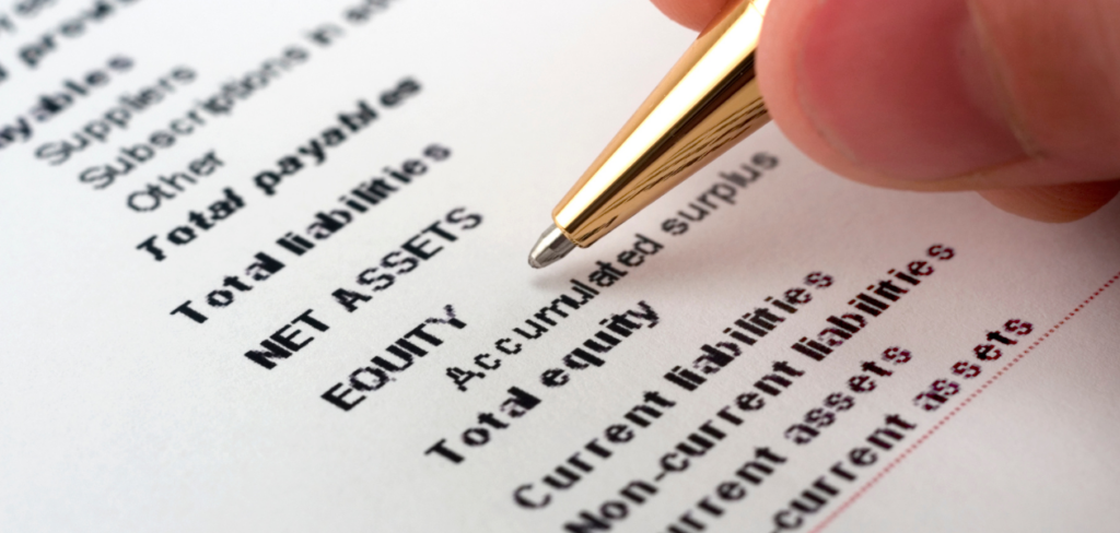 A close up of a hand holding a pen in front of a document of assets and liabilities.