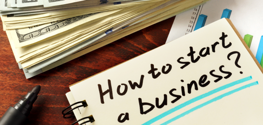 A notepad with 'How to Start a Business?' written on it rests on a wooden table, next to a stack of cash.