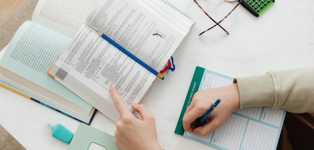 Top view of hands writing on a book with a pen on a table with a notebook, pen, glasses, and a cup of coffee.