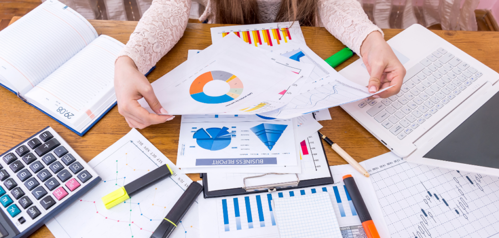 A person is sitting at a desk with a laptop and papers, comparing business graphs and data.
