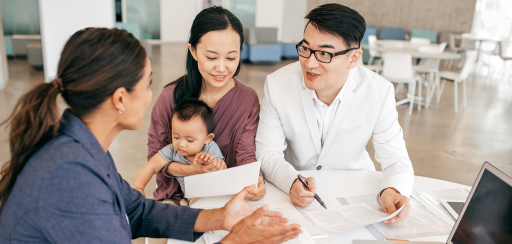 A husband is holding papers, and his wife is holding their kid while talking to a financial advisor.