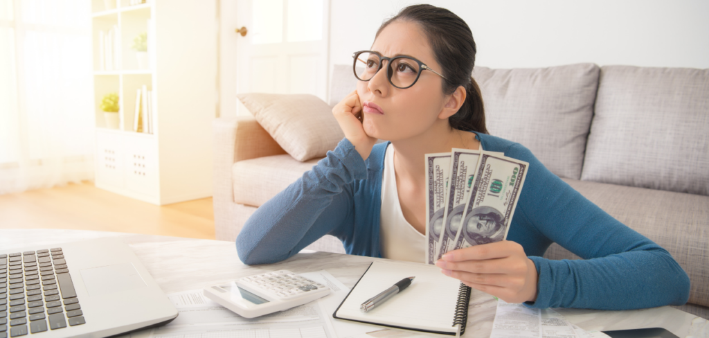 A woman is thinking while holding cash, with a pen, notebook, and laptop in her living room.
