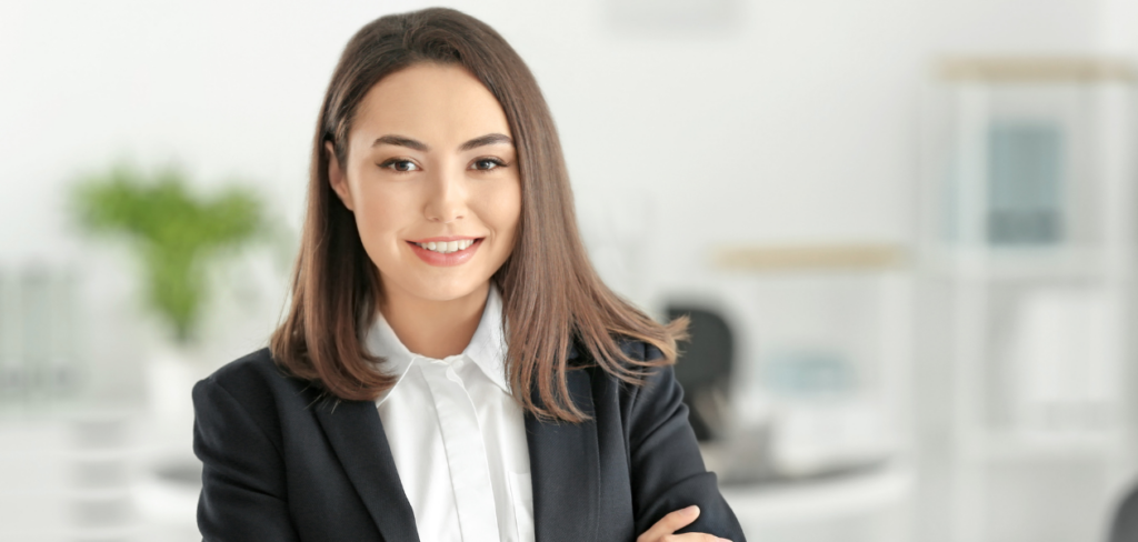 A woman in a business suit is standing with her arms crossed, smiling.