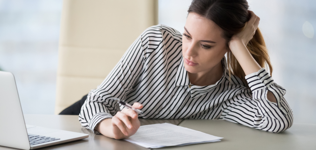 A woman feeling the lack of motivation sitting at a desk with a laptop, pen, and paper.