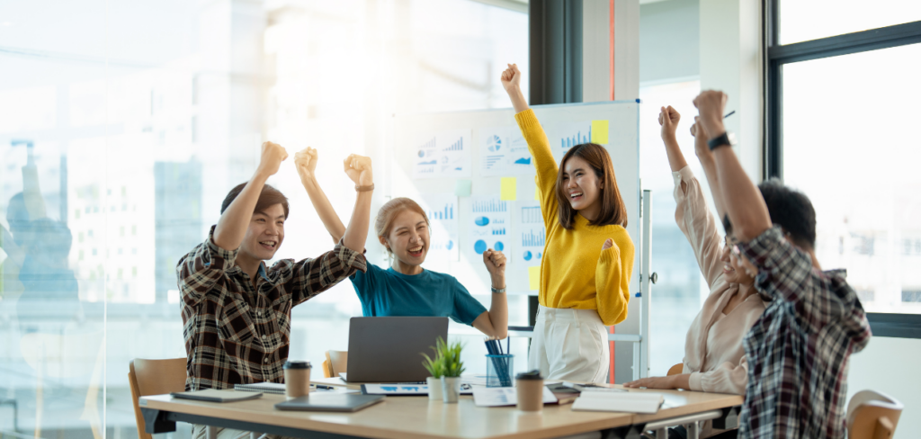 A group of people is seated around a table, raising their hands in the air to celebrate their success.
