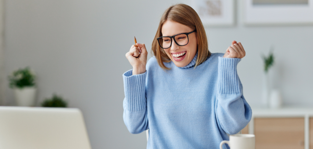 A happy young woman laughing and yelling in excitement after reading a message on her laptop while sitting at a table at home.
