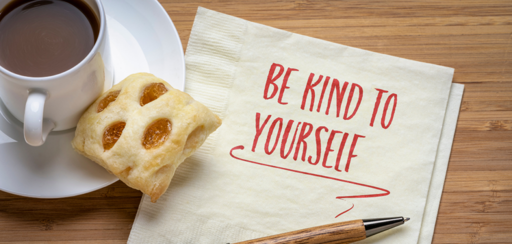 Handwritten message 'Be kind to yourself' on a napkin, with coffee, pastry, and a pen.