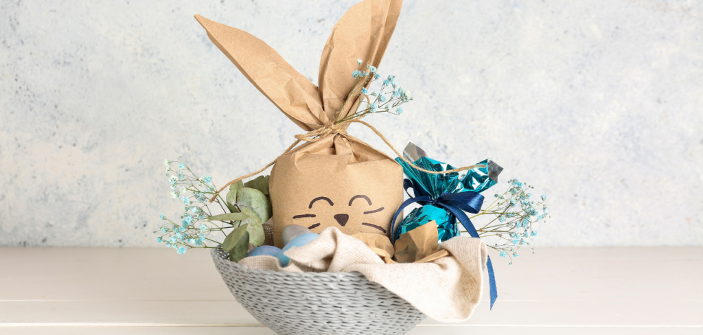 Basket with an Easter bunny gift bag, Easter eggs, and flowers on a white wooden table.