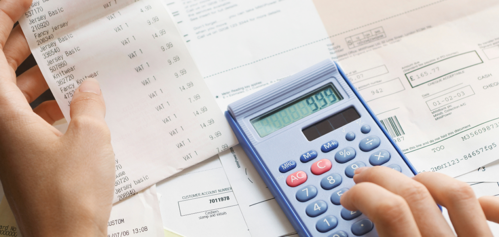 A hand using a calculator to tally the total expenses from receipts.
