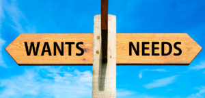 Wooden signpost with two arrows pointing in opposite directions labeled 'WANTS' and ‘NEEDS'.