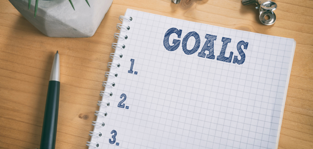 List of goals on a notepad placed alongside a pen, clips, and a plant on a wooden desk.